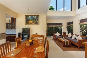 A spacious dining area with a wooden table set for six, surrounded by wooden chairs. A large painting adorns the wall above the table. - Kadiga Villas - 3 Bedroom Villa (Bali)