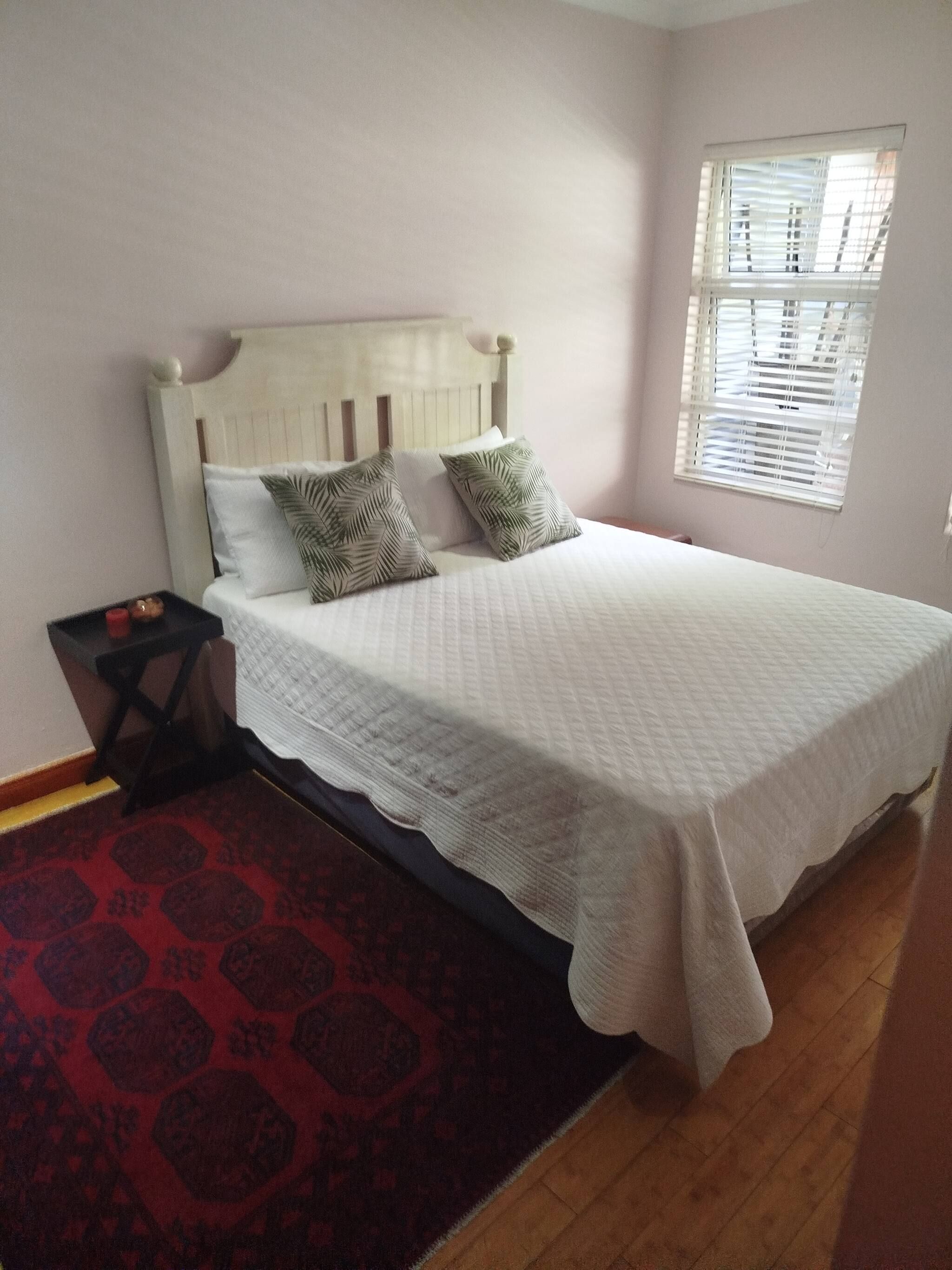 Una cama con cabecera blanca, dos almohadas, un tapete rojo estampado y una ventana con persianas.