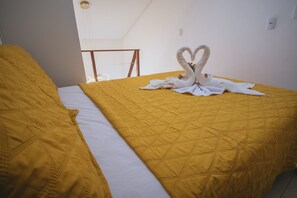 A hotel room with a bed featuring a yellow quilt and white pillows, a white swan towel decoration, and a view of the sky through a window. - Bella V. Ap. 304 C - Andres Sardi (Ipojuca)