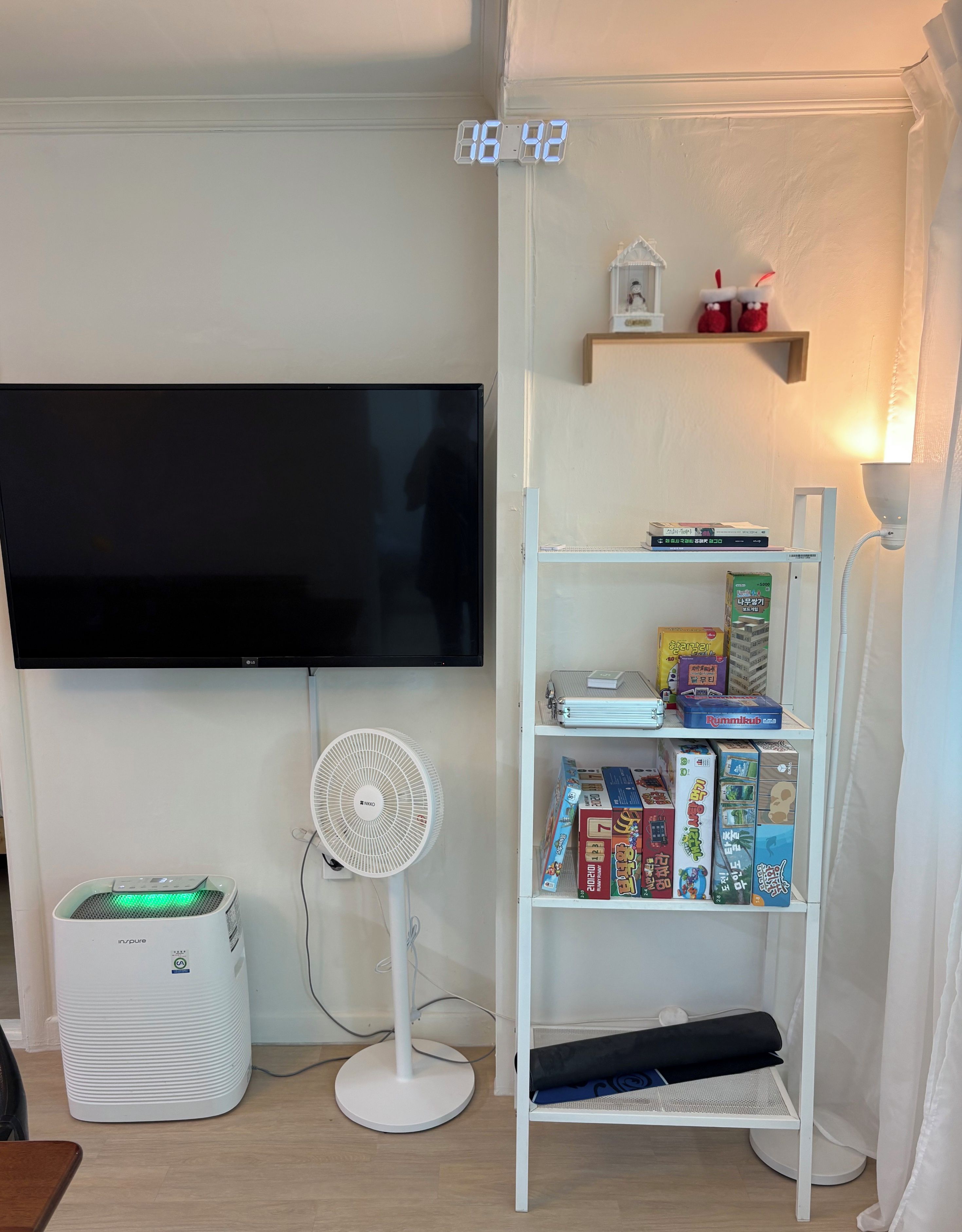 Ein Zimmer mit einem an der Wand befestigten Flachbildfernseher, einem Standventilator, einem weißen Heizgerät und einem Bücherregal mit verschiedenen Gegenständen.