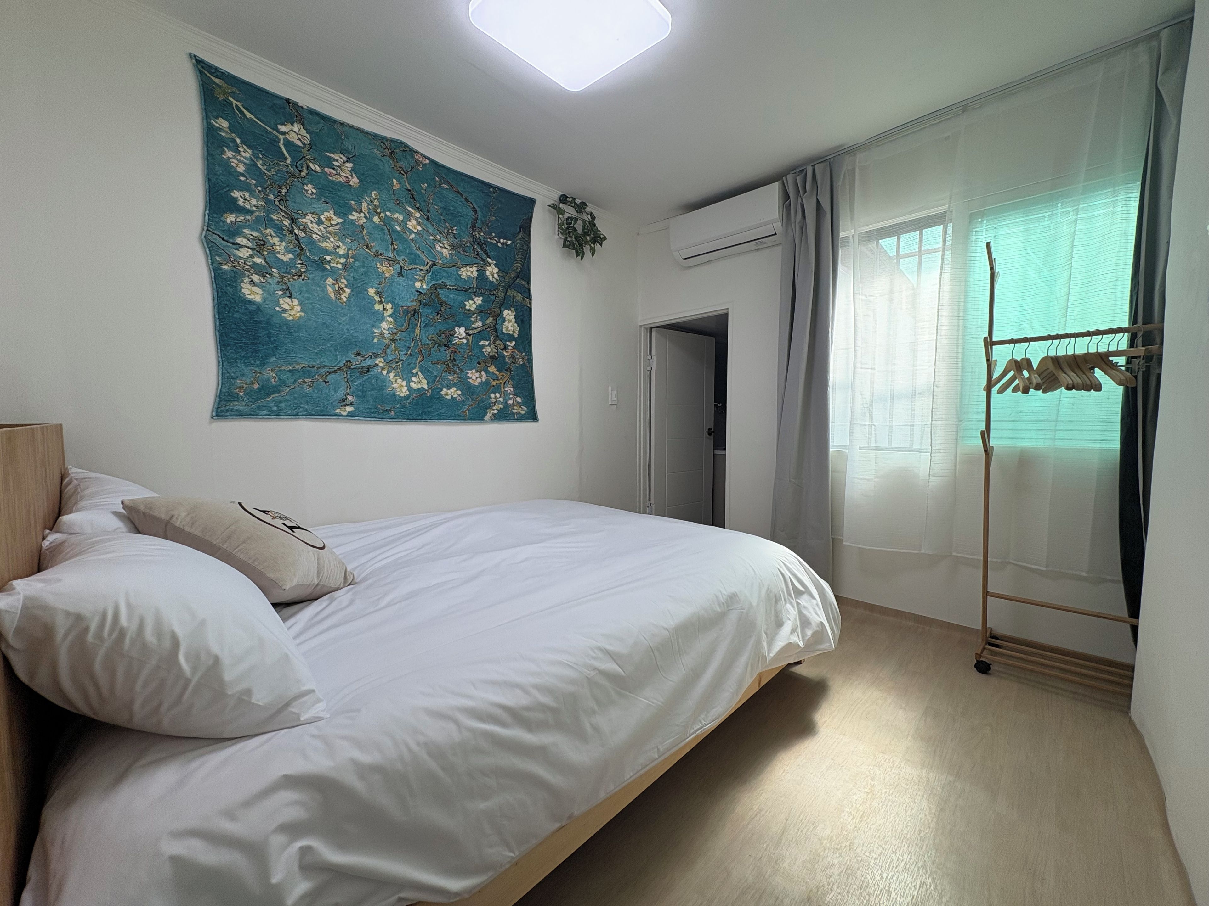 A bedroom with a bed, a large floral tapestry, an air conditioning unit, and a clothing rack.
