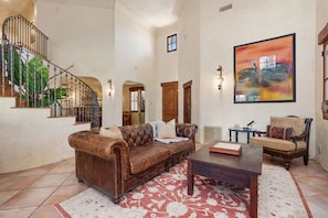 A spacious living room with a brown leather sofa, a wooden coffee table, and a red patterned rug. A staircase with a metal railing is visible in the background. - Mediterranean style retreat. (Solana Beach)