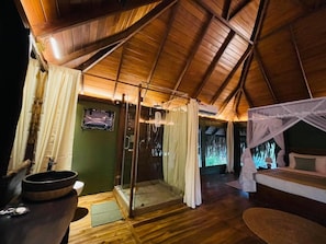 Een kamer met een houten plafond, een douchecabine van glas, een bed met hemel en een houten tafel met een kom.