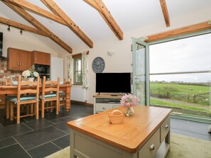 A dining area with a wooden table and chairs, a television, and a view of a green field through a glass door. - Ash Cottage (Truro)