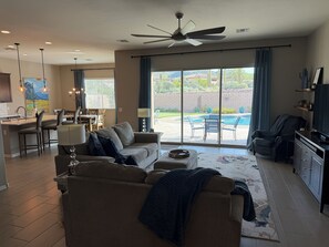 Ein geräumiges Wohnzimmer mit einer Couch, einem Couchtisch und einem Fernseher. Es gibt einen Essbereich mit Stühlen und einem Tisch, sowie eine Küche mit einer Arbeitsfläche und Pendelleuchten.