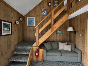 Un intérieur en bois avec un escalier, un canapé gris et des éléments de décoration murale, notamment une affiche du Vermont et une enseigne publicitaire pour la marque de vêtements Corinthia.