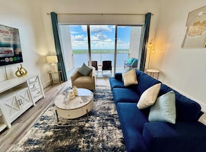 Ein Wohnzimmer mit einer Couch, Couchtisch und einem Fernseher auf einem Standfuß. Es gibt einen Balkon mit Blick auf den Himmel und Wolken.