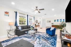 A modern living room with a sofa, armchair, and coffee table, a ceiling fan, and a large window with curtains. - The Beachside Escape I New Pool & Spa! (Hollywood)