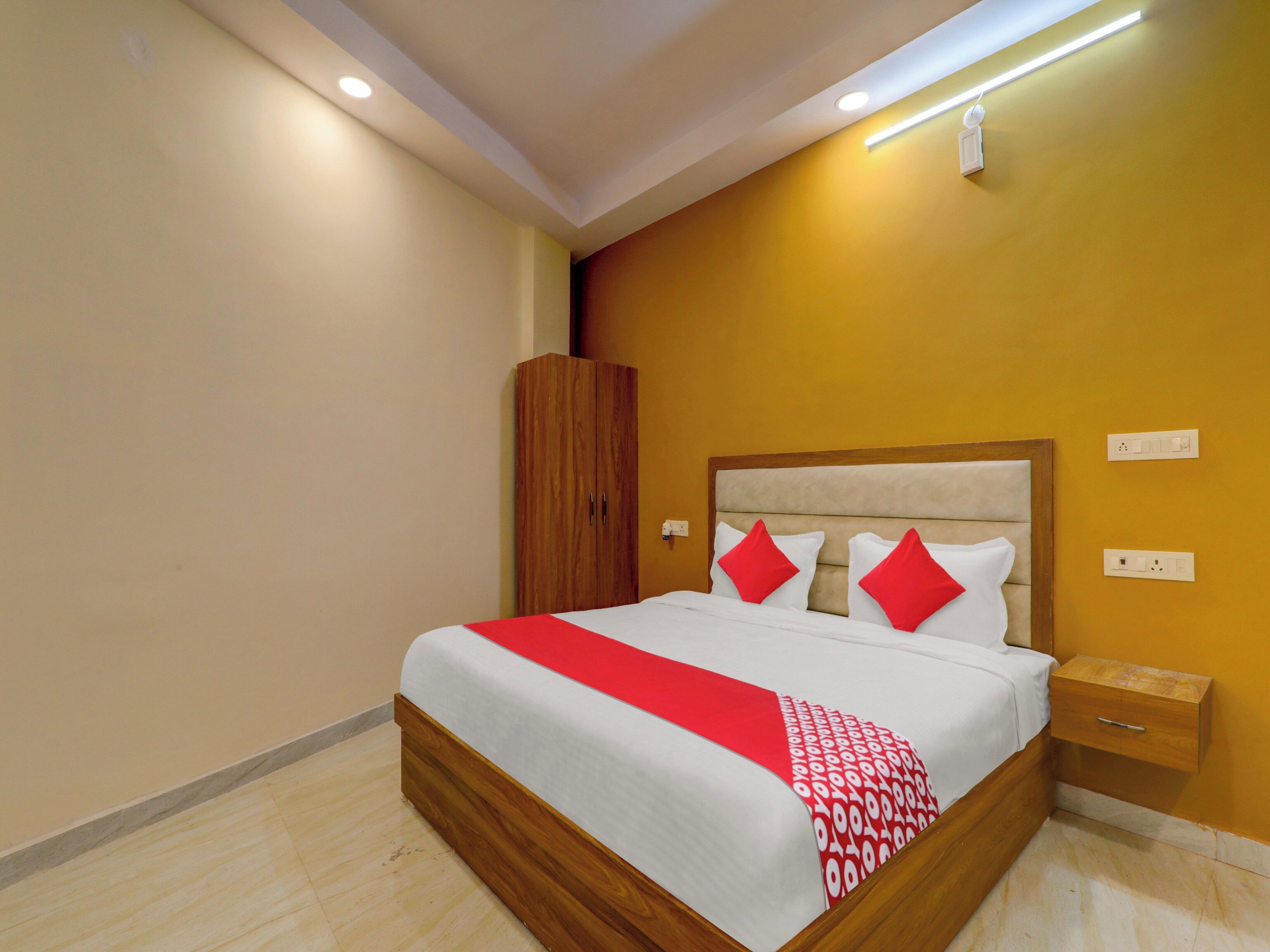 Een net opgemaakt bed met een rood-wit sprei, een houten garderobe en een nachtkastje met een lade.