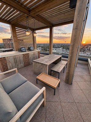 Un patio exterior con mobiliario de madera, una parrilla y vistas a la ciudad al atardecer.
