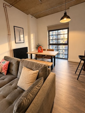 A modern living room with a sectional sofa, a dining table, and a view of a cityscape through large windows. - Luxury 2BR Wembley Loft | Skyline Views | Near Wembley Stadium (Wembley)