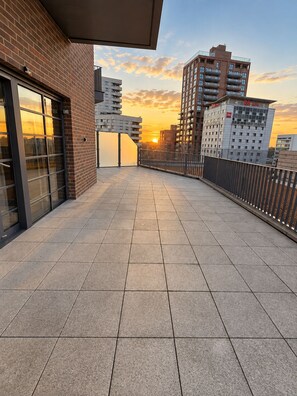 Een dakterras met uitzicht op de zonsondergang, een bakstenen gebouw en moderne hoogbouw op de achtergrond.
