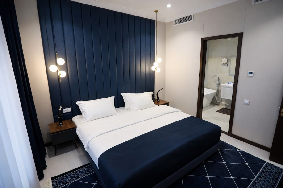A hotel room with a large bed, bedside tables, a mirror, and a bathroom visible through an open door.
