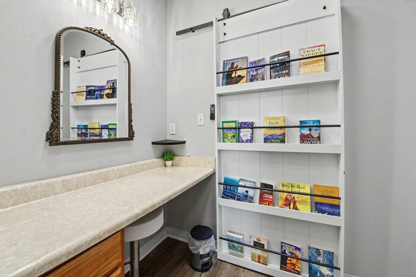 Une salle de bain avec un meuble-lavabo blanc, un miroir doté d’un cadre orné, une étagère avec des livres et une poubelle.