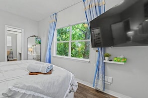 A bedroom with a bed, a television, a window with curtains, and a small shelf with items. - Cottage by The Sea, Family Retreat w/ Game Room (Biloxi)