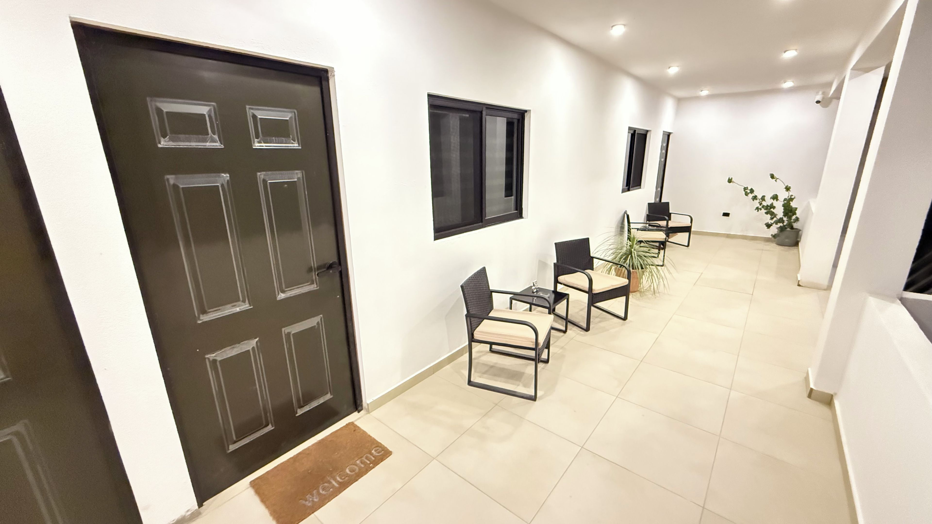 A hallway with a dark door, a welcome mat, and a row of chairs with a potted plant.