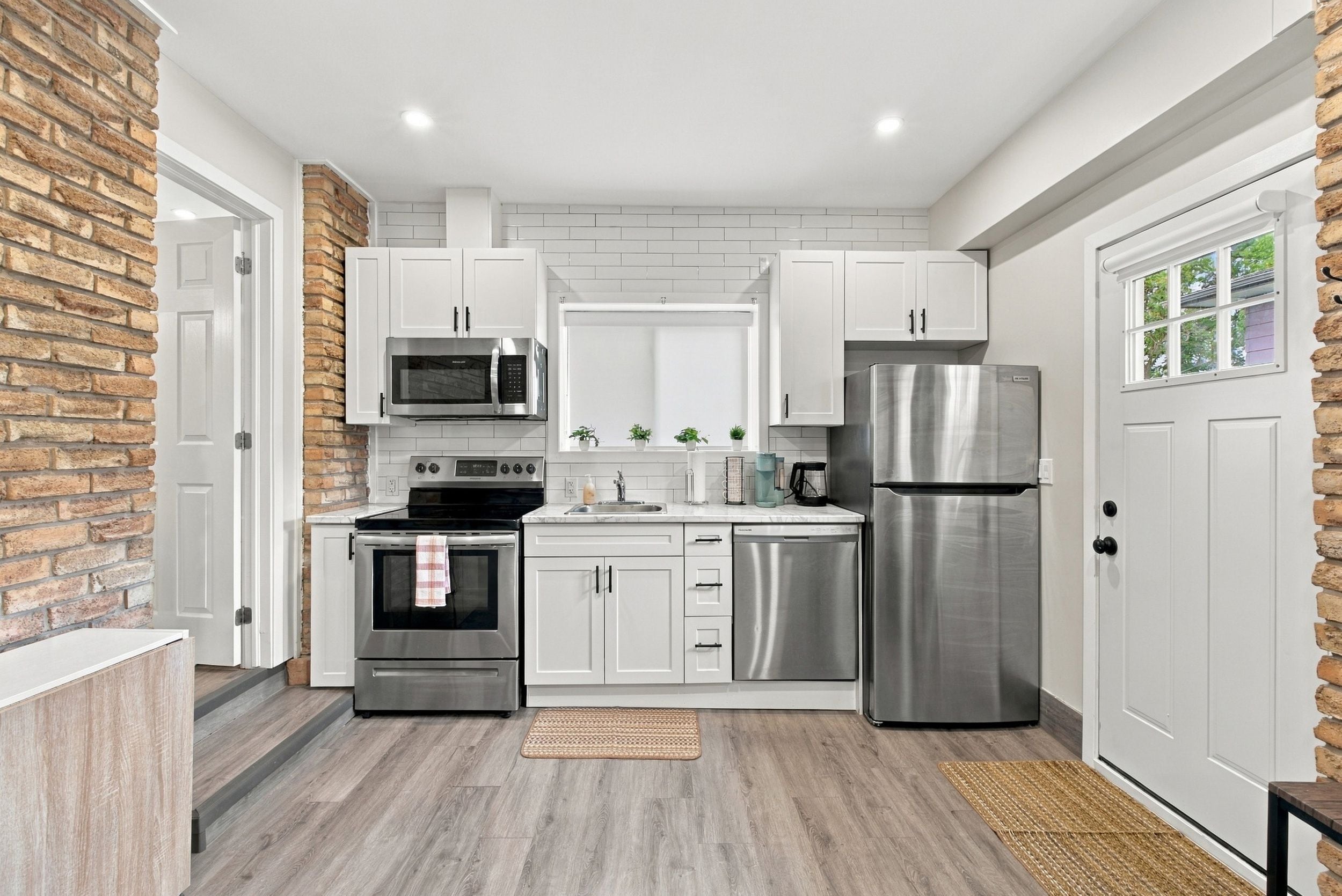 A modern kitchen with white cabinets, stainless steel appliances, a brick accent wall, and a wooden floor.