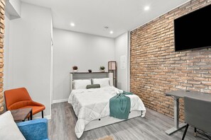 Un dormitorio moderno con pared de ladrillo decorativa, cama con ropa blanca, televisor montado en la pared y una mesita.