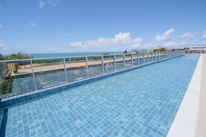 A long swimming pool with clear blue water, surrounded by a glass railing, and overlooking a beach and ocean. - Nixxus 216 (Ipojuca)