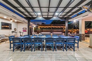 An outdoor dining area with a long table and chairs, surrounded by a covered patio with string lights. - Sunset Villa | Pool ·BBQ ·Outdoor kitchen & Grill (Hialeah)