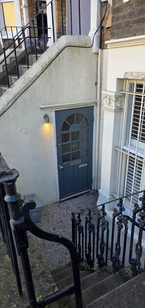 Una escalera conduce a una puerta con reja de metal, una lámpara de pared y una ventana con persianas.