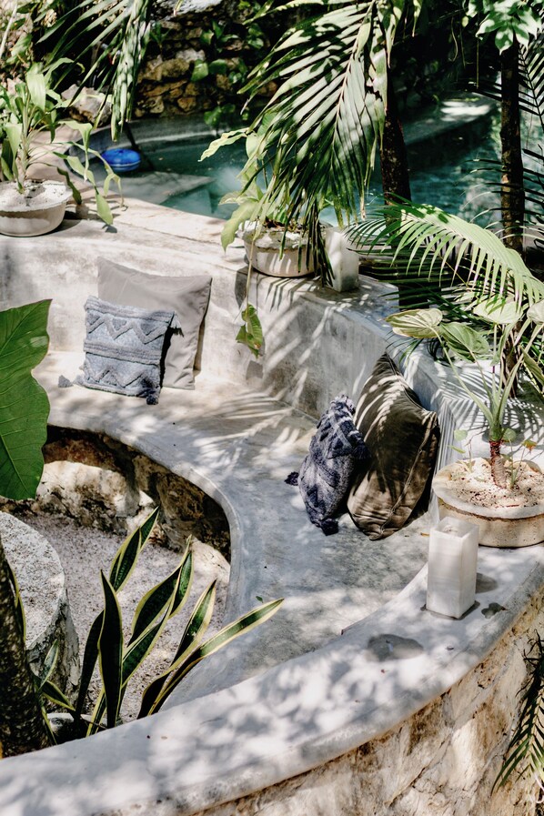 A stone bench with cushions and a small plant, surrounded by greenery and a stone wall.