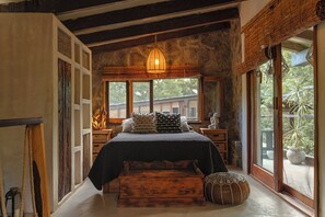 A bedroom with a large bed, wooden beams, a hanging light fixture, and a view of greenery outside.