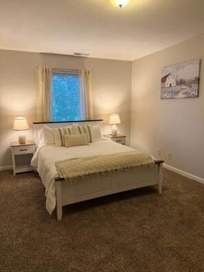 A bedroom with a bed, two bedside tables, a window with curtains, a wall-mounted painting, and carpeted flooring.