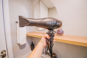 A hand holding a hairdryer with a cord, in a bathroom with a towel hanging on the wall and a wooden shelf with a decorative item. - Nixxus 316 (Ipojuca)
