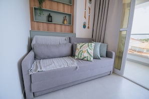 A grey sofa with a textured throw blanket and a tropical leaf pillow, against a wall with a wooden panel and a hanging decoration. - Nixxus 316 (Ipojuca)