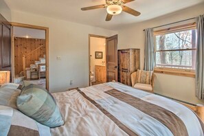 A bedroom with a large bed, a ceiling fan, a window with curtains, and a wooden door leading to a staircase. - Woodsy WV Cabin near Lost River w Deck & Fire Pit! (Mathias)