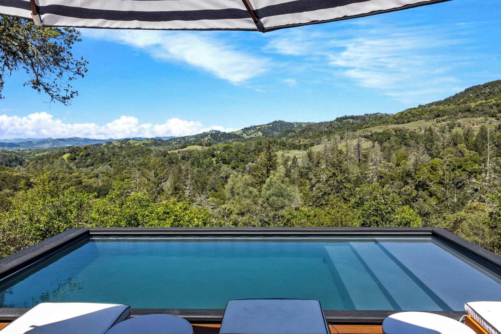 Blick auf einen Swimmingpool mit Holzterrasse und zwei weißen Liegestühlen, mit Blick auf eine Berglandschaft.