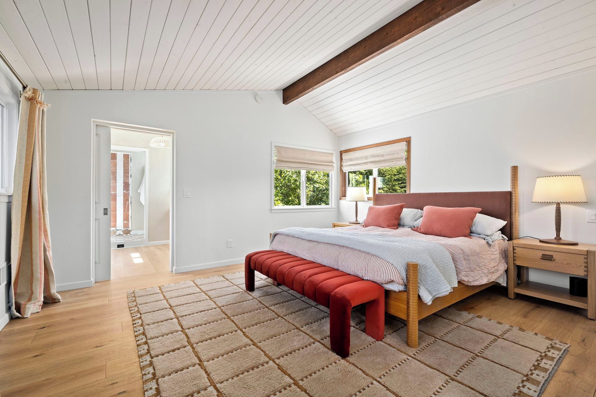 Quarto com teto de madeira, cama com cabeceira, mesa de cabeceira, abajur e um banco.