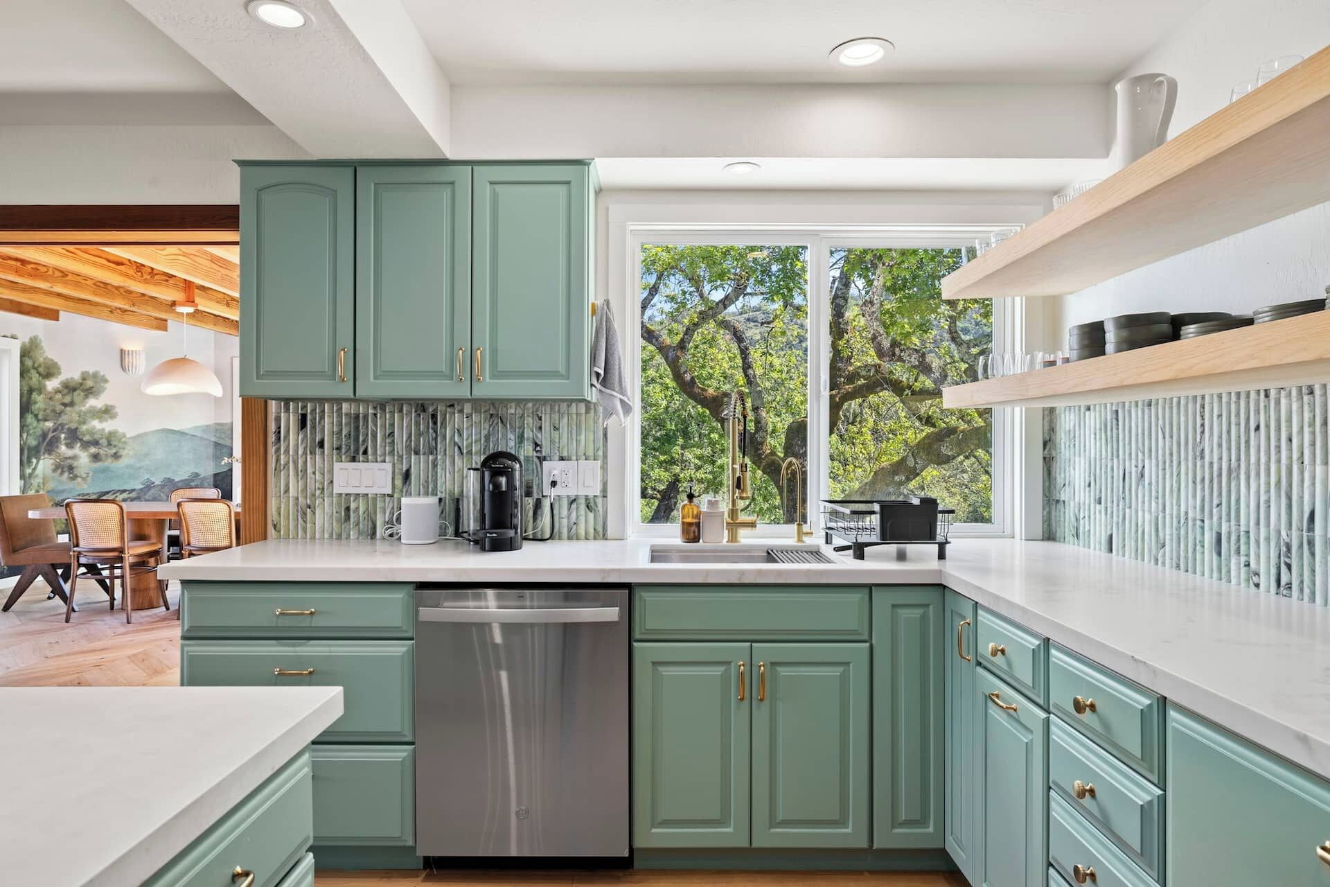 A modern kitchen with teal cabinetry, a stainless steel dishwasher, and a large window offering a view of trees.
