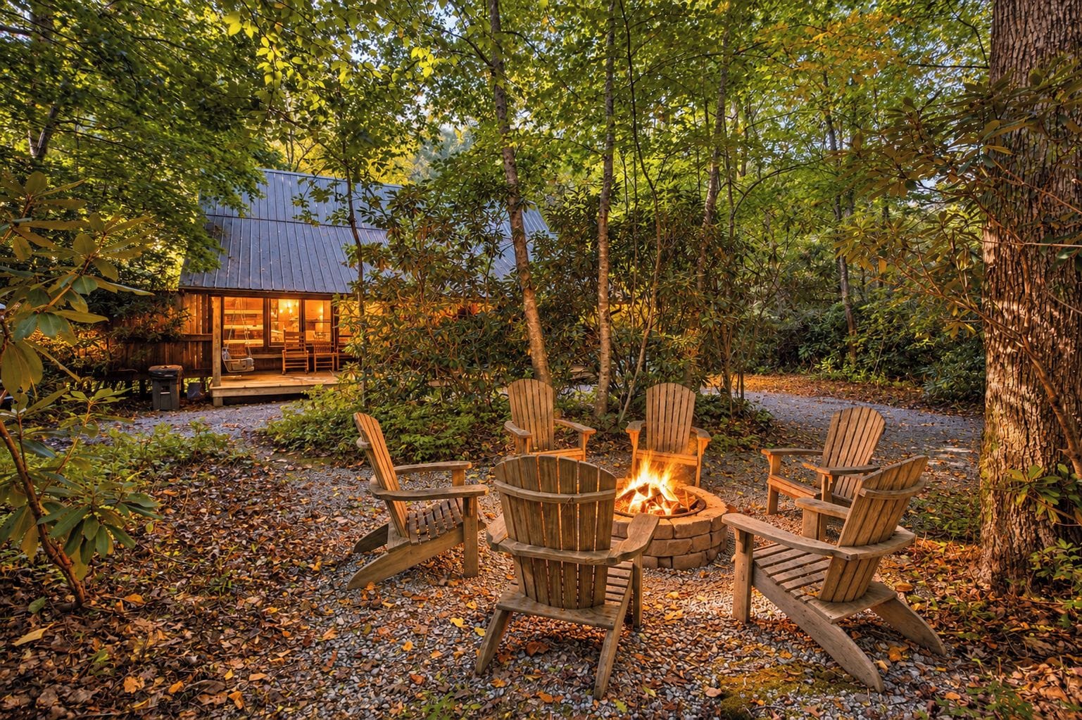 A cozy outdoor seating area with a fire pit and Adirondack chairs, surrounded by trees and a cabin in the background.