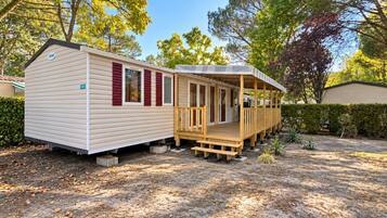 A mobile home with a porch and a small deck, surrounded by trees and a hedge.