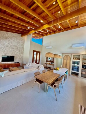 A modern living room with a wooden ceiling, a large flat-screen TV, a sofa, a dining table with chairs, and a kitchen area with an oven and microwave. - Amore'mare | Charm, Comfort and Sea (Ubatuba)