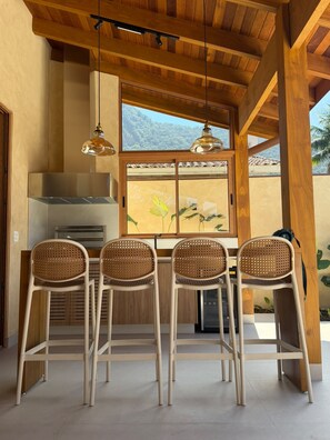 A dining area with four rattan chairs, a stainless steel range hood, and a large window with a view of greenery. - Amore'mare | Charm, Comfort and Sea (Ubatuba)