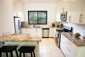 A modern kitchen with white cabinets, a granite countertop, stainless steel appliances, and a window with blinds. - Walking distance to everything, the beach, and town | Tropical condo (Coco)