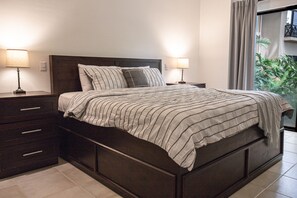 A bed with a striped bedspread, two bedside lamps, a dark headboard, and a view of greenery through a window. - Walking distance to everything, the beach, and town | Tropical condo (Coco)