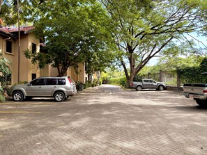 Rua residencial com carros estacionados, árvores e um edifício com telhado em telhas.