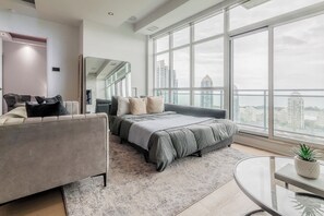 A modern bedroom with a large window, a bed, a sofa, and a small table with a plant. - One-Of-A-Kind Parklawn Penthouse! Unreal Lake View | Parking Available (Toronto)