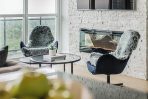 Uma sala de estar moderna com lareira, duas poltronas aconchegantes, uma mesa de centro redonda e vista para o verde lá fora.