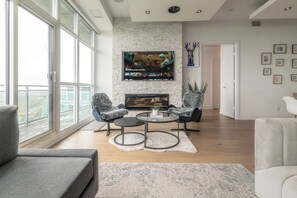 A modern living room with a fireplace, a flat-screen TV mounted on a brick wall, and a large window offering a city view. - One-Of-A-Kind Parklawn Penthouse! Unreal Lake View | Parking Available (Toronto)