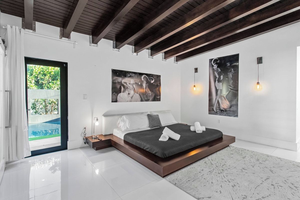 A modern bedroom with a large bed, a view of a swimming pool, and artwork on the walls.