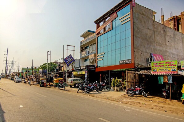 A street with various shops and a building with a large glass facade. - Hotel O Sky In Near By Sector 62 (Ghaziabad)