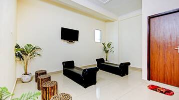 Un área de recepción con un televisor de pantalla plana montado en la pared, dos sofás negros, una mesa de centro de madera y una planta en maceta.