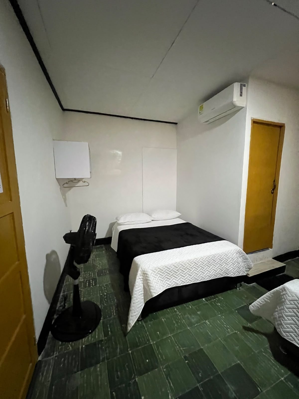 Una habitación pequeña con cama individual, suelo verde, aire acondicionado blanco empotrado en la pared, puerta de madera y un ventilador negro.