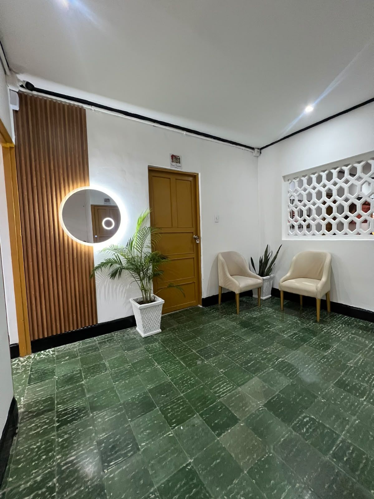 Un área de recepción con un panel de pared de madera, un espejo redondo, dos sillas, una planta en maceta y un panel decorativo.