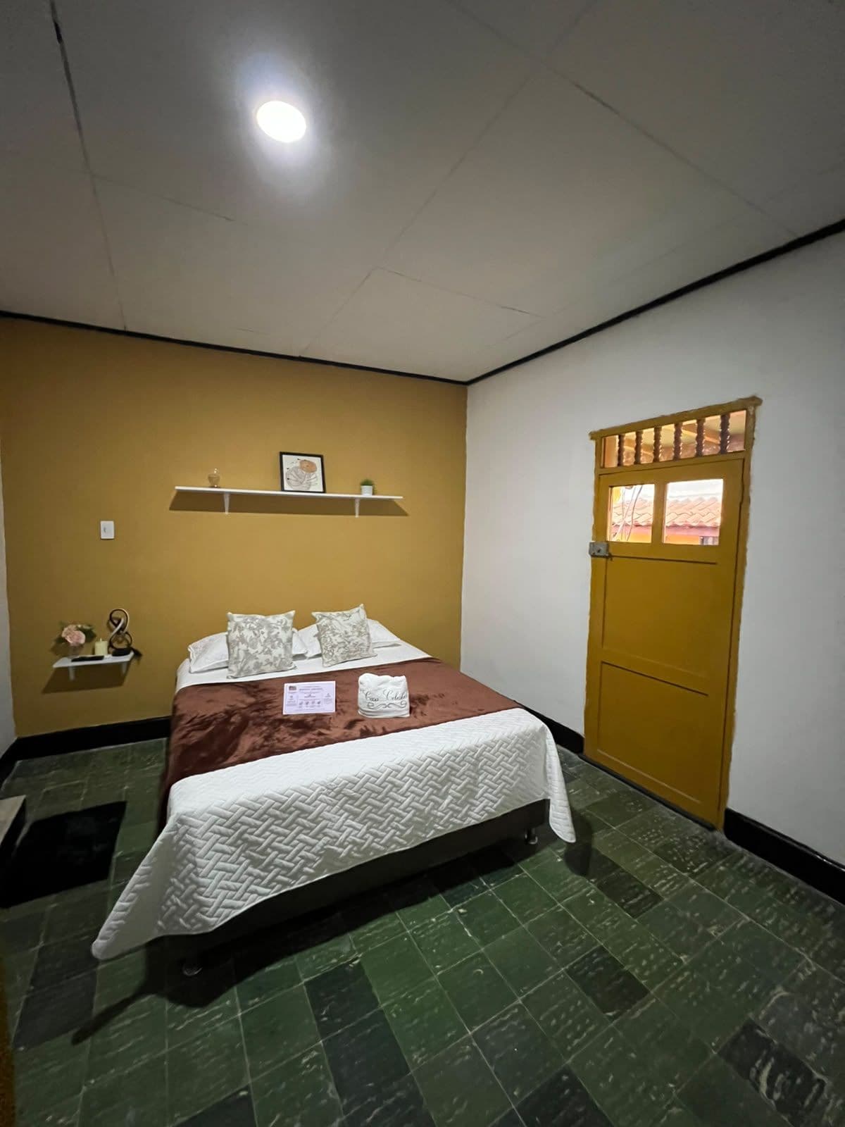 A hotel room with a bed, a small shelf with decorative items, a door with a window, and a tiled floor.